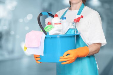 A cleaning lady with a bucket and cleaning products stands on a blurred background.