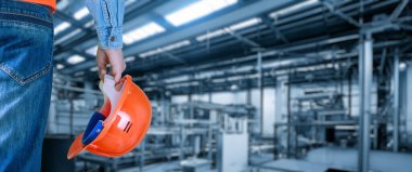 Elinde miğfer olan bir işçi üretim tesisinin arka planında duruyor..