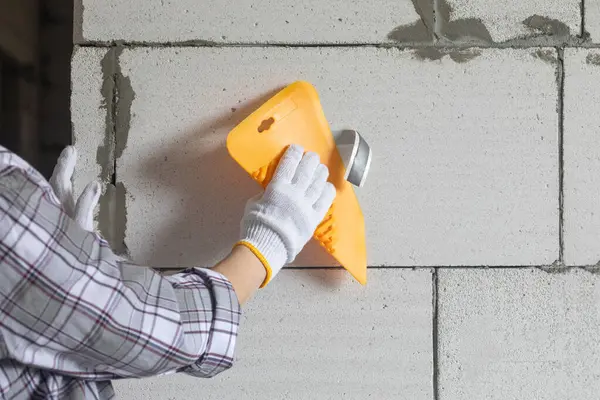 A worker applies construction adhesive tape to a wall using a plastic putty knife.