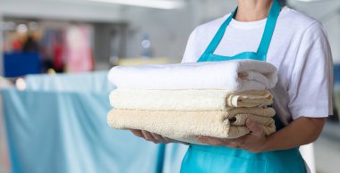 Mavi önlüklü temizlikçi kadın arka planda bulanık havlular tutuyor..