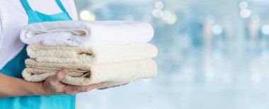 Mavi önlüklü temizlikçi kadın arka planda temiz havlu yığını tutuyor..