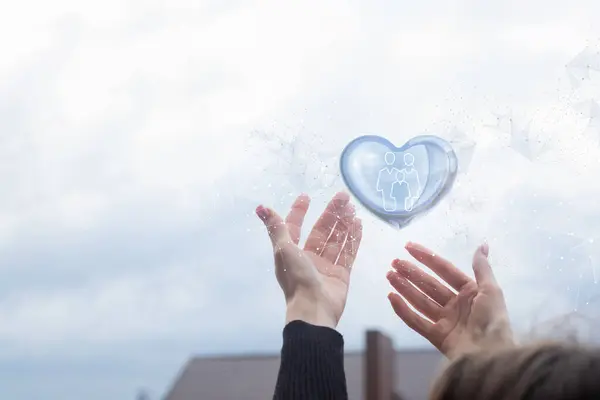 stock image The concept of digital security and family care. Hands support a heart with a family icon in it against a sky background.