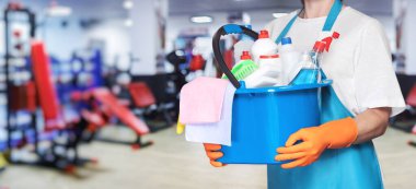 Spor salonunda temizlik ve hijyen. Elinde kovayla temizlik malzemesi olan bir temizlikçi kadın spor salonunun arka planında..