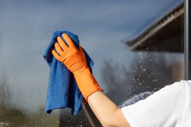Uzlaşmasız temizlik mükemmel pencereler. Beyaz tişörtlü ve turuncu eldivenli bir temizlikçinin camı mavi bir mikrofiber bezle iyice temizlediği pencere temizleme işlemi..