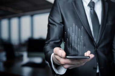 Resmi giyinmiş bir iş adamı, cihazdan çıkan dinamik grafik verileri gösteren bir akıllı telefon bulundurur..