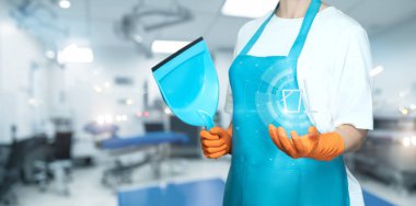 Önlüklü bir temizlikçi hastane ortamında bir kepçe ve dijital cihaz tutuyor. Bireysel olarak tıbbi çevredeki temizliği korumaya odaklanmıştır..