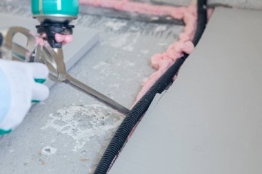 Bir kişi köpük aplikatörü kullanarak zemin yalıtım kanalının etrafına köpük yerleştiriyor. Köpük, inşaat sırasında yalıtım etkisini artırmak için yüzeylere yapışır..