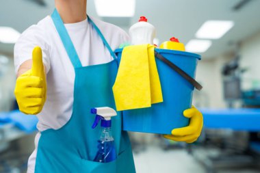 Eldivenli bir işçi, malzeme dolu bir temizlik kovasını tutarken baş parmağını kaldırır. Bir tıbbi tesisin temiz çevresi hijyen ve güvenlik standartlarını artırır.