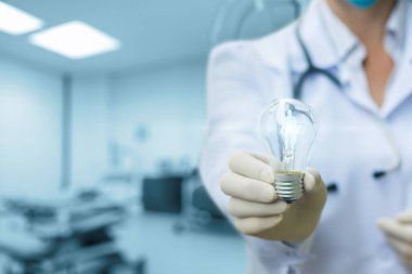 A healthcare professional holds a light bulb in a modern examination room, symbolizing innovative ideas in patient care and diagnostics. The background shows medical equipment.