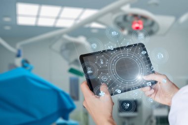 Bir tıp uzmanı, ameliyat odasında dijital ikonları gösteren bir tabletle cerrahi verimliliği ve hasta bakımını arttıran bir etkileşim içindedir. Çevre modern ve yüksek teknolojili..