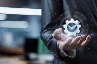 Takım elbiseli bir kişi, modern ofis ortamında verimliliği ve ilerlemeyi simgeleyen işaretli parlak bir vites sembolü taşıyor..