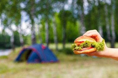 Yeşil ağaçların altındaki bir nehrin kenarında kamp yaparken, elinde marul ve domates olan taze yapılmış bir hamburger. Arka planda renkli bir çadır kurulmuş. Açık hava atmosferini arttırıyor..
