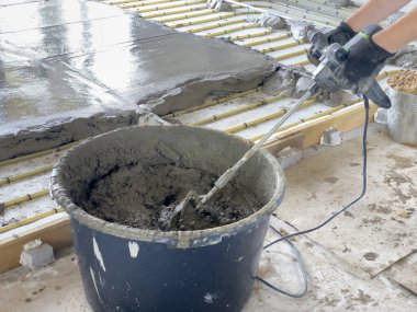 İşçiler beton döşeme projesi için bir karıştırma aracı ve büyük bir kova kullanarak çimentoyu karıştırıyorlar. Düzen, istikrar için ahşap kalasları olan bir inşaat alanı..