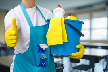 Neşeli temizlik malzemeleri, parlak sınıf ortamında takım çalışması. Bir ekip üyesi verimli bir güne hazırlanırken bir temizlik kovası ve erzak sergiliyor..