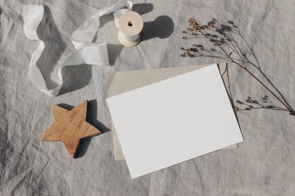 Christmas stationery. Blank greeting card, invitation mockup. Wooden star ornament, craft envelope and dry grass in sunlight. Beige linen tablecloth. White ribbon, winter wedding flat lay, top