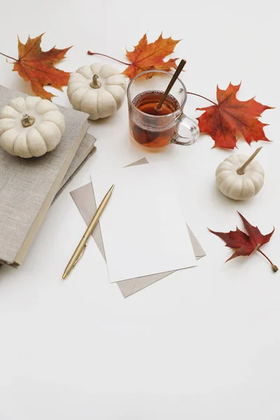 Fall still life. Autumn stationery. Blank greeting card, invitation mockup. Pumpkins, red maple leaves isolated on white table background. Breakfast scene. Glass cup of tea, golden pen and book, top