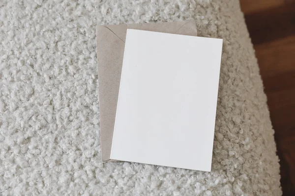 Neutral stationery still life scene. Blank greeting card, invitation mockup and craft envelope on white boucle sofa upholstery. Teddy textured fabric, blurred oak wooden parquet floor. Top view.