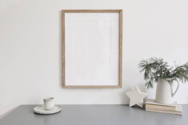 Noel hala hayattır. Boş ahşap çerçeve modeli. Bir fincan kahve, gri masadaki sürahi içinde çam ağacı dalları. Eski kitaplar, seramik beyaz yıldız dekoru, İskandinav şenlik posterleri kışlık ev dekorasyonu..