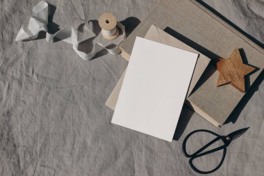 Noel kırtasiyesi. Boş tebrik kartı, davetiye maketi. Tahta yıldız süsü, eski kitaplar ve güneş ışığında beyaz ipek kurdele. Bej keten masa örtüsü. Siyah antika makas, kış düğünü düzlüğü.