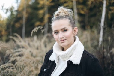 Güzel, uzun saçlı, beyaz saçlı, beyaz örgü süveterli, siyah ceketli bir kızın hayat tarzı portresi. Kuru çim çayırı. Autmn, sonbahar manzarası, sarı huş ağaçları, bulanık ağaçlar..