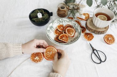 Noel çelengi yapan kadınların eli yakındır. Bir fincan çay, portakal, meyve ve çam, beyaz keten masa örtüsü üzerinde okaliptüs ağacı dalları, şenlikli kış sanatları masası. Seçici odak.