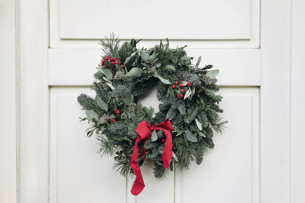 Winter holidays festive still live. Advent wreath made of green fir, pine tree, eucalyptus and olive branches. Red rose hips berries with silk ribbon, White wooden front door background, home decor.