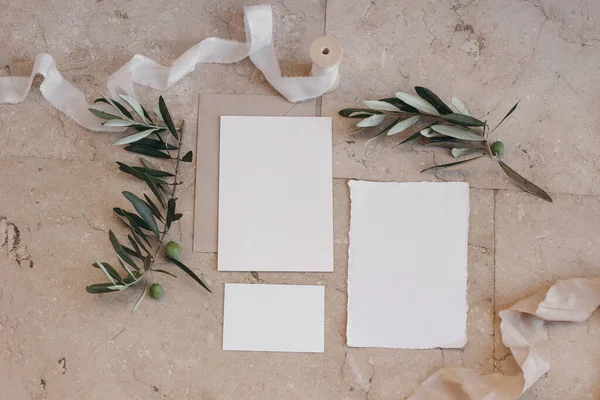 Summer wedding stationery set, suite. Italian, Greek mock-up scene with blank paper greeting cards, envelope on beige marble tiles background. Green olive tree branches, silk ribbon, flat lay, top.