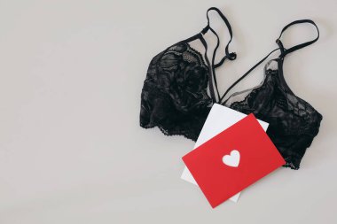 Black lace bra and red postcard, invitation mockup with heart on beige table background. Women sexy underwear, lingerie. Gift Idea for Womens Day, Saint Valentines holiday, copy space, flatlay, top