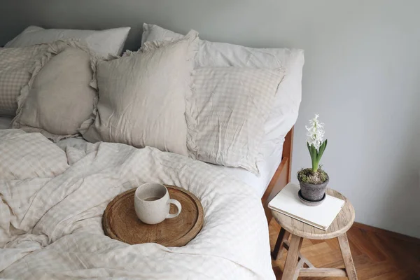 Breakfast in bed concept. Cup of coffee on wooden tray. Bedroom view. Checkered beige pillows, blanket. Vintage hocker, night stand. White hyacinth in flower pot, blurred elegant spring interior.