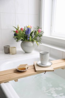 Cup of coffee, candles, book and soap on wooden board. Elegant Scandinavian bathroom interior with white tiles.Blurred background. Colorful tulips, hyacinth spring bouquet, bath with foam. Vertical.