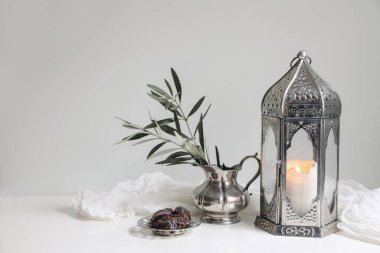 Ramadan Kareem still life. Glowing ornamental lantern. Olive tree branches in silver jug, vase. Date fruit, silver tray. Iftar dinner. Eid mubarak, Eid ul Fitr holiday celebration, festive background. clipart