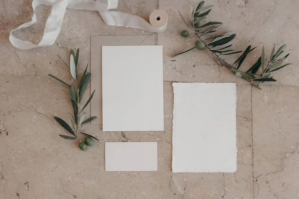 Italian summer wedding stationery set. Mock-up scene with blank paper greeting, RSVP cards on beige marble tiles background. Green olive tree branches, silk ribbonMediterranean flat lay, top view.