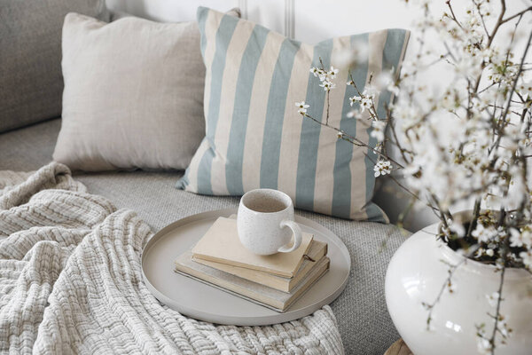 Весенний натюрморт. Чашка кофе, чай на стопке книг. Круглый бежевый стол. Цветущие вишневые ветви сливы в керамической вазе. Уютный постельный диван, подушки, размытый фон. Интерьер дома. Топ