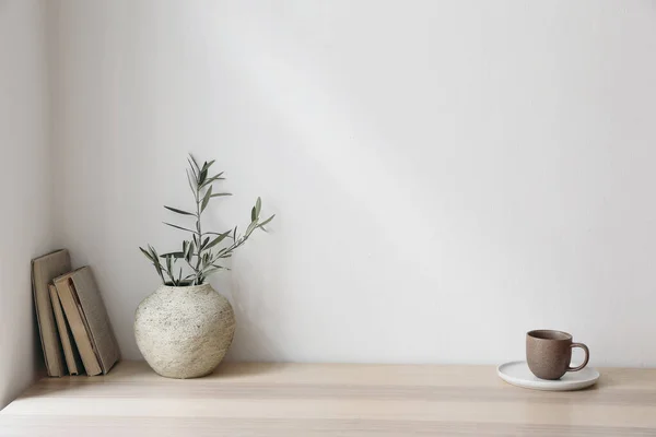 stock image Olive tree branches in beige textured vase. Brown cup of coffee, tea and old books on wooden table. White wall background. Minimalistic Scandinavian interior, dinning room, summer breakfast.