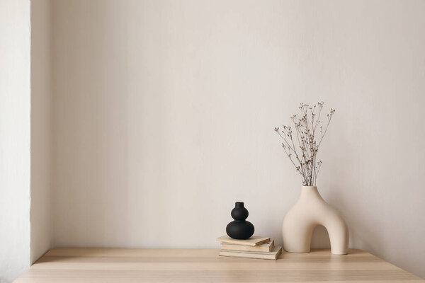 Artistic interior still life. Modern organic, geometric shaped sculptures. Vase with dry flowers, grass and old books on wooden table. Home staging, minimal decor concept, empty beige wall background.