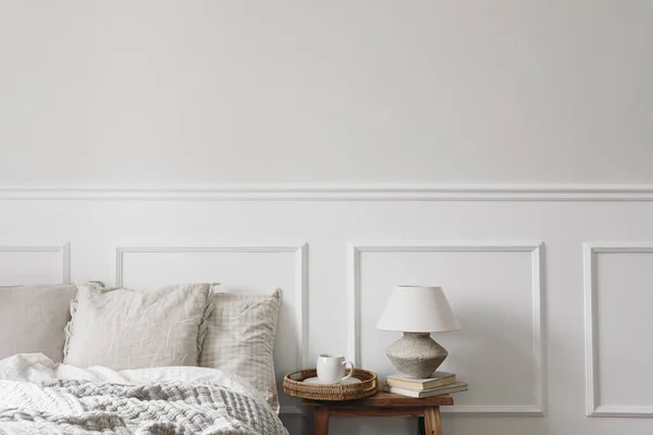 Cup of coffee on wicker tray. Textured clay table lamp, old books on vintage wooden bench, night stand. Bedroom view. Checkered beige pillows, linen blanket, elegant Scandinavian interior, copy space.