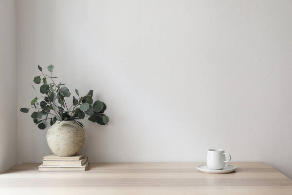 Minimal breakfast still life. Cup of coffee, tea. Empty white wall in sunlight table. Textured vase with silver eucalyptus leaves, branches on old books, elegant Scandinavian working space, office
