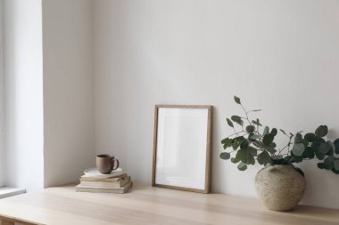Bir fincan kahve, kitaplar. Ahşap masa, masa üzerinde boş dikey çerçeve modeli. Gümüş okaliptüs dallı vazo. Asgari çalışma alanı, ev ofisi, İskandinav iç tasarımı. Yan görünüm.