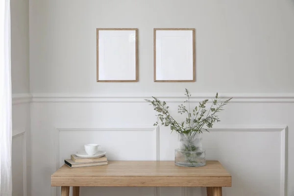 Two empty vertical picture frame mockups hanging on wall. Cup of coffee, books. Wooden desk, table. Vase, green grasses and cow parsley. Minimal working space, home office, elegant Scandi interior.