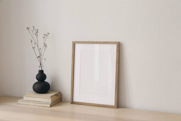 Black organic shaped vase with dry flowers, grass on old books. Blank picture frame mockup, wooden table at home. Minimal Scandi boho interior. Beige wall background. Empty copy space, side view.
