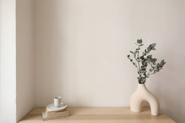 Modern organik vazoda okaliptüs ağacı dalı. Bir fincan kahve, ahşap masada eski kitaplar. Boş bej duvar modeli, kopyalama alanı. Zarif oturma odası, İskandinav minimalist iç mekan, ev, insan yok..