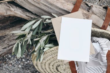 Yaz yaşam tarzı kompozisyonu. Boş tebrik kartı davetiyesi maketi. Fransız sepeti, zeytin dalı dallı saman torbası, havlu: Akdeniz tasarımı, yanlama tahtası ve kaygan çakıl plajı arka planı.