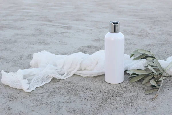 White cosmetic plastic bottle mockup with green olive tree branch and muslin veil. Natural liquid soap, shampoo or moisturizer. Grey grunge concrete floor background. Summer skin care. Cosmetology.
