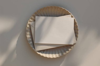 Minimum yaz düğün kırtasiyesi. Bej zarflı boş yatay tebrik kartı modeli, davetiye. Güneş ışığında seramik deniz tarağı tabağı. Gölgeli masa arkaplanı, düz, üst