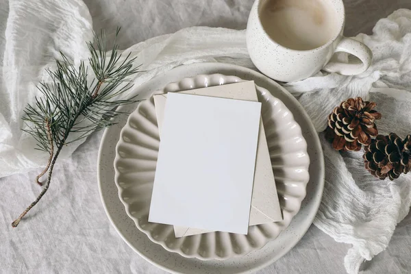Christmas still life. Blank greeting card, invitation mockup on ceramic plate. Pine cones and pine tree branches on white linen tablecloth. Cup of coffee, winter breakfast, festive holiday flat lay