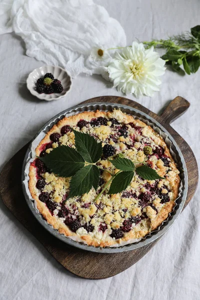 Lifestyle food, kitchen still life. Homemade sweet blackberry pie, tart. Old wooden chopping board. Blurred bowl with berries with dahlia, cosmos flowers. Grey linen table cloth. Breakfast, top view.