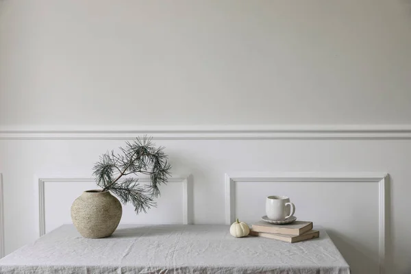 Elegant Christmas interior. Winter dinner table setting. Cup of coffee, old books and little white pumpkin. Pine tree branches in vintage vase. White wall background, classic moulding trim, no people