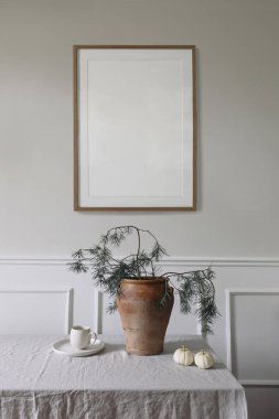 Zarif bir iç mekan, Noel sofrası dekorasyonu. Kış sanat kompozisyonu. Boş ahşap çerçeve bej duvarda asılı. Vazoda çam ağacı dalları. Bir fincan kahve, beyaz balkabağı, dikey..