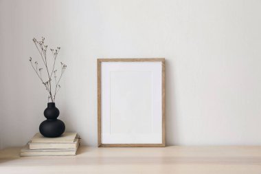 Kuru çiçekli siyah organik vazo, eski kitaplardaki çimenler. Boş dikey ahşap çerçeve modeli. Ofis masası. Minimal Scandi boho iç mekanı. Beyaz duvar arkaplanı, boş telif alanı.