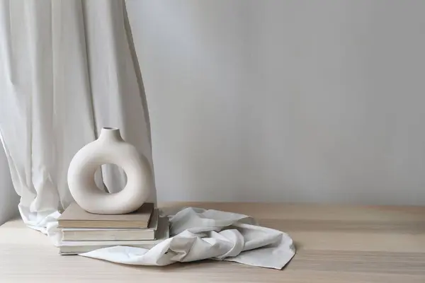 Modern organic shaped vase on old books, beige table. Linen curtain, folded drapery and empty white wall background. Aesthetic sunlight shadows, minimalist template for home room interior product.